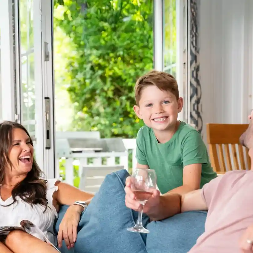 A woman and a man sit on a couch, laughing, while a boy stands between them smiling cheerfully. The background features large windows with greenery outside, creating a bright and inviting atmosphere.