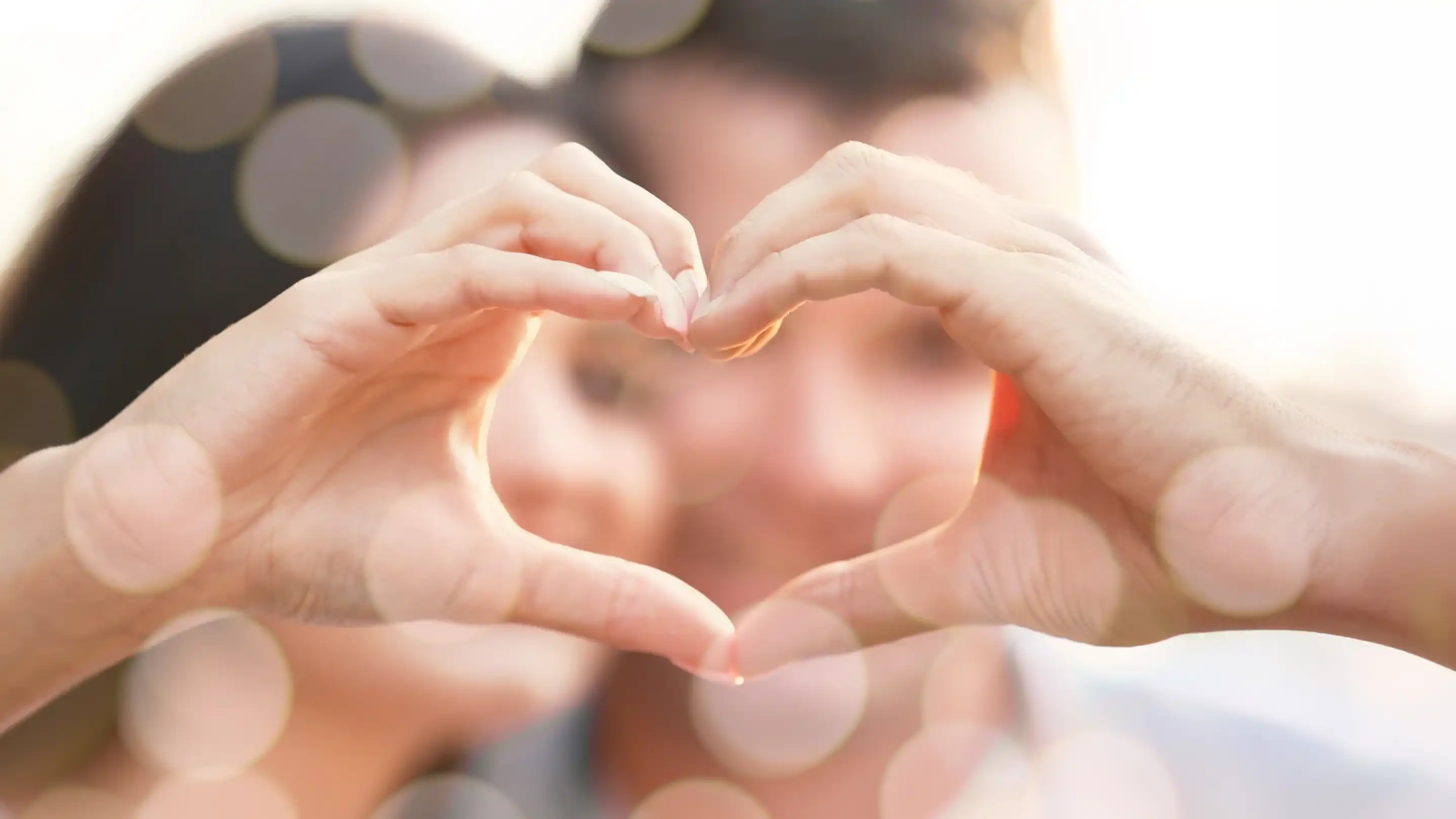 Two hands create a heart shape in the foreground, with a couple's smiling faces softly blurred in the background. Bokeh lights add a dreamy effect to the scene.