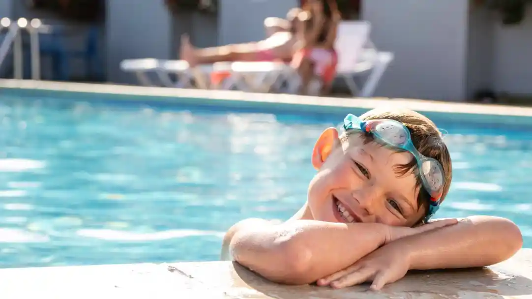 A smiling boy with swim goggles rests his arms on the edge of a pool, enjoying a sunny day. In the background, a woman relaxes on a lounger.
