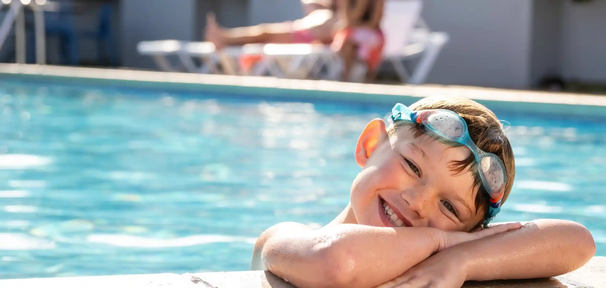 A smiling boy with swim goggles rests his arms on the edge of a pool, enjoying a sunny day. In the background, a woman relaxes on a lounger.