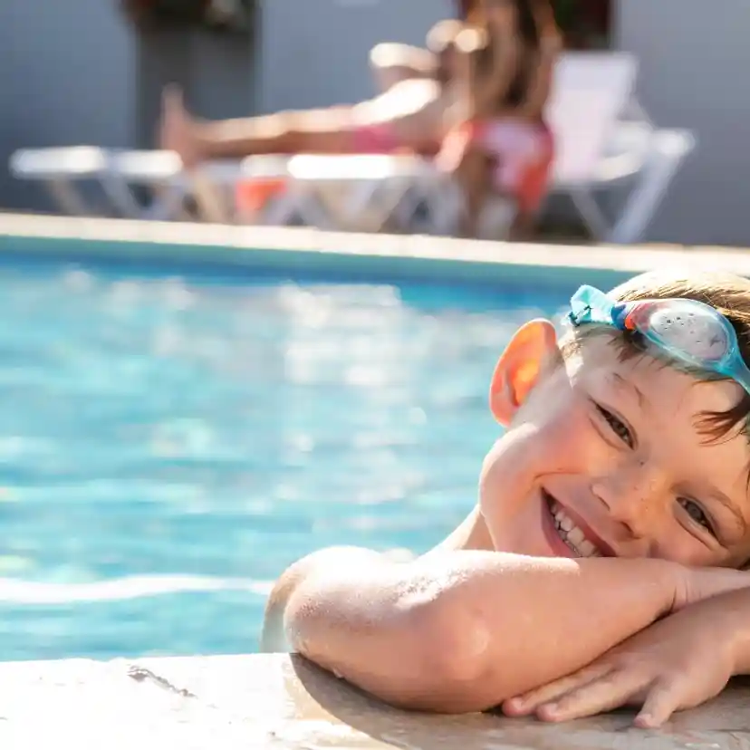 A smiling boy with swim goggles rests his arms on the edge of a pool, enjoying a sunny day. In the background, a woman relaxes on a lounger.