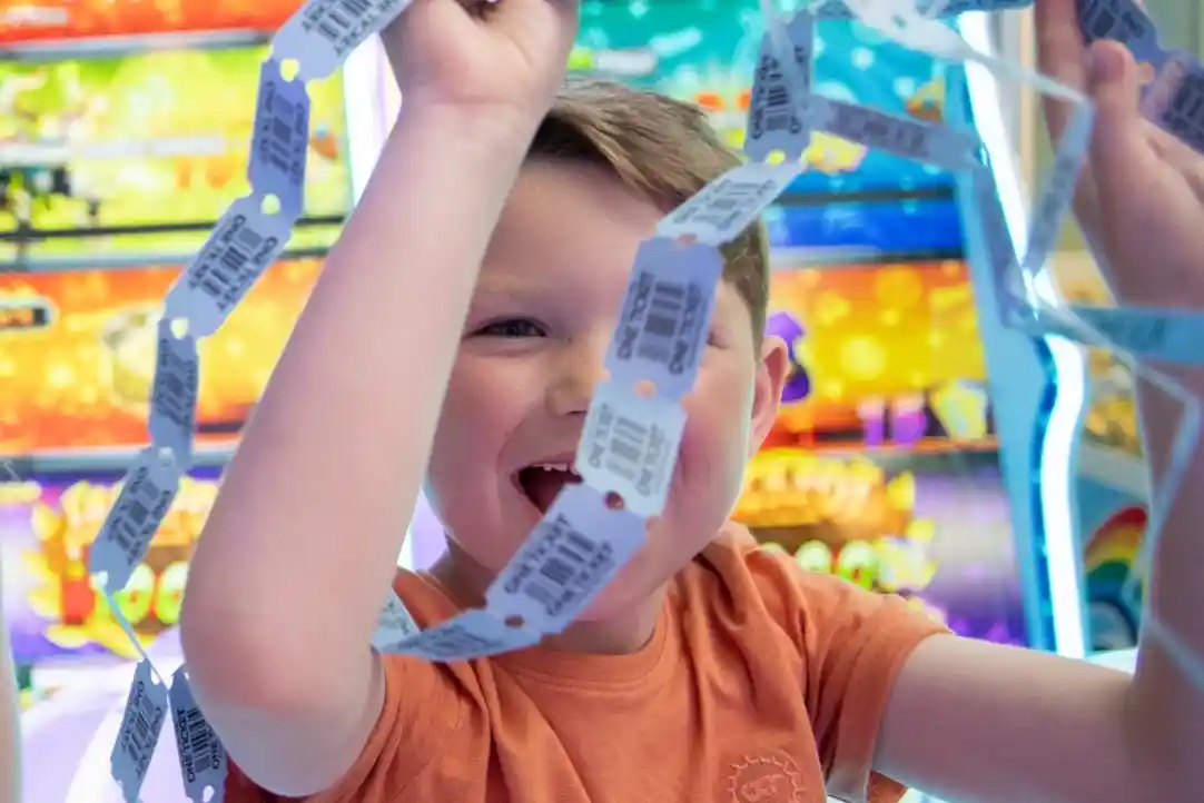 A young boy with a joyful expression holds up a chain of arcade tickets, surrounded by colorful arcade games in the background.