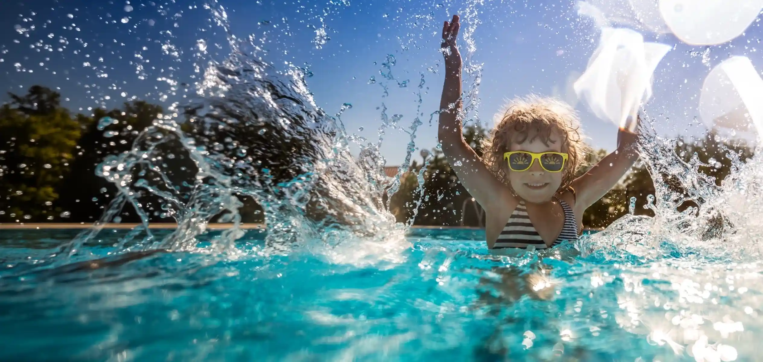 A child wearing yellow sunglasses joyfully splashes in a pool, with sunlight reflecting off the water. Trees are visible in the background under a clear blue sky.