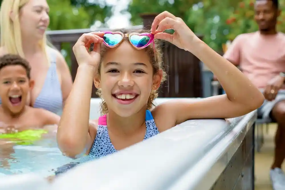 A smiling girl with curly hair holds up heart-shaped sunglasses while sitting in a spa or pool. Behind her, a woman and a boy are enjoying the water, and a man is seated nearby.