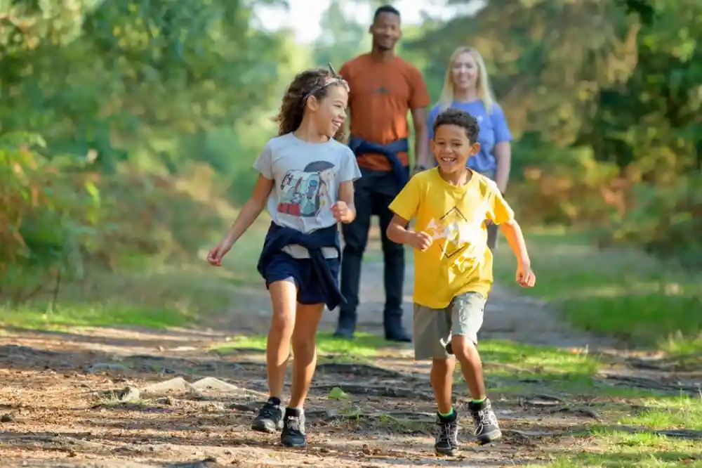 Two children joyfully run along a forest trail, surrounded by greenery. The girl wears a gray shirt and the boy is in a yellow shirt. In the background, two adults smile as they walk together.