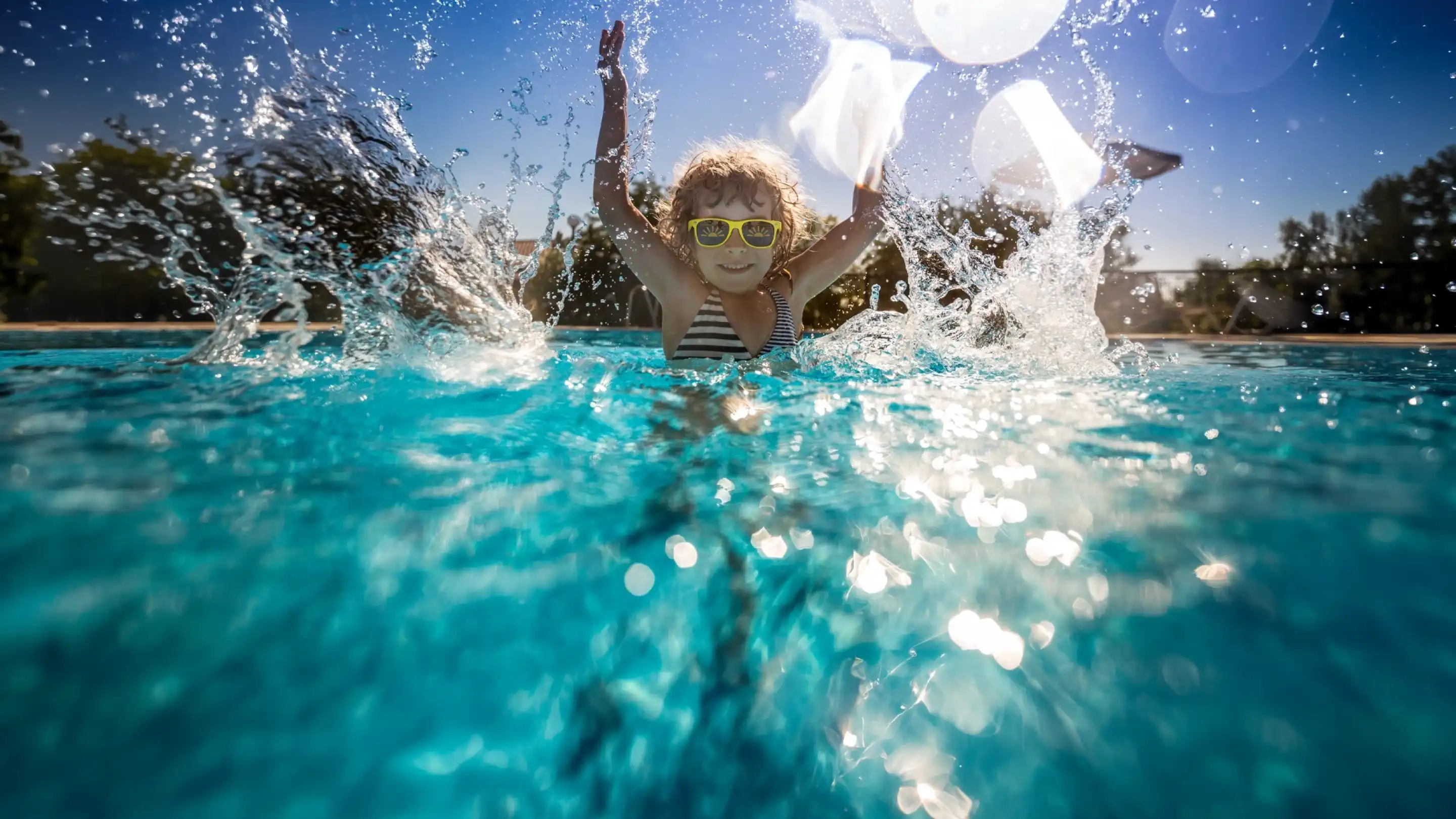 A joyful child wearing sunglasses splashes water while playing in a swimming pool on a sunny day. Sunlight glimmers on the surface of the water, creating a vibrant and cheerful atmosphere.