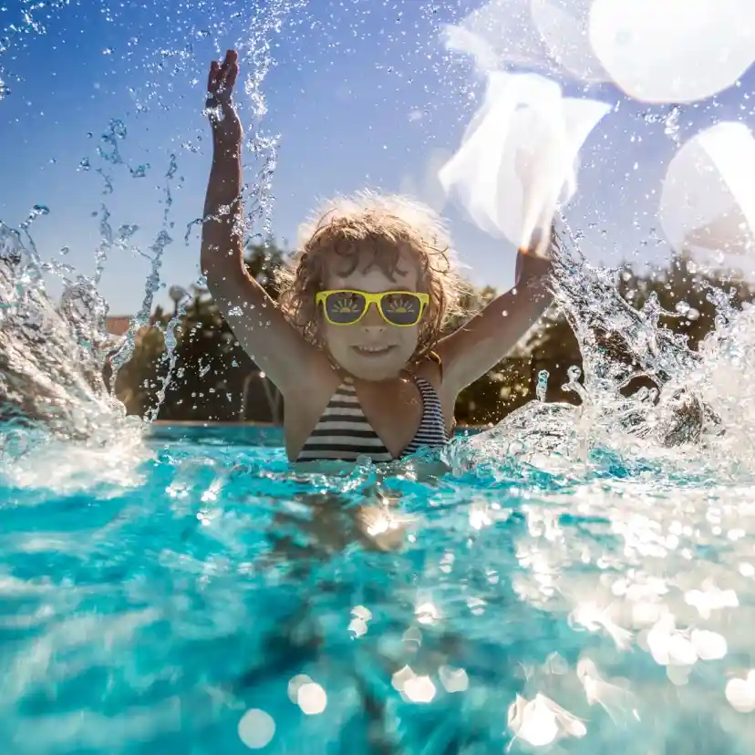 A joyful child wearing sunglasses splashes in a clear blue pool under a bright sunny sky, raising their arms in excitement. Water droplets sparkle around them.