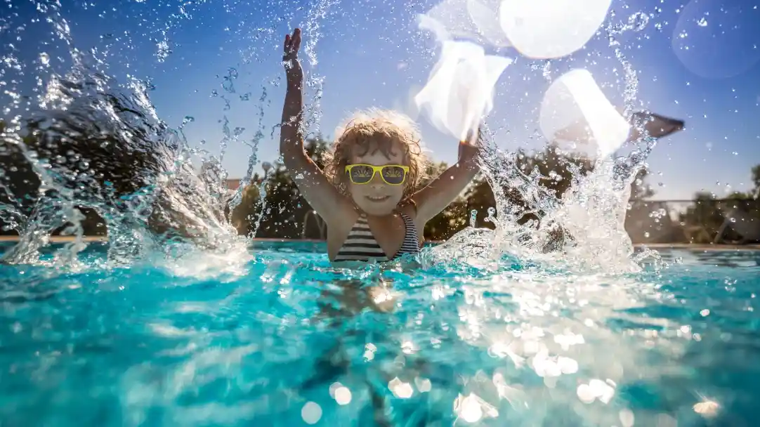 A joyful child wearing sunglasses splashes in a clear blue pool under a bright sunny sky, raising their arms in excitement. Water droplets sparkle around them.