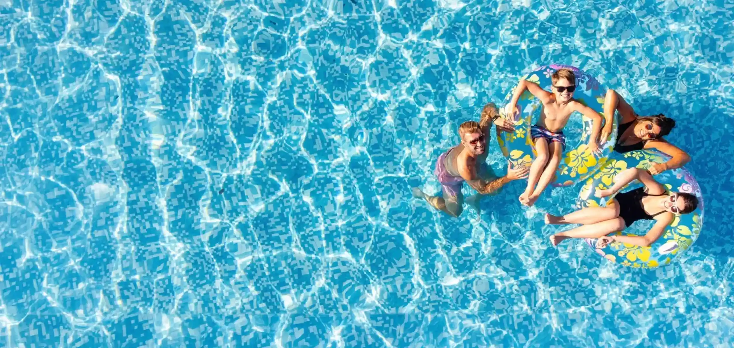Aerial view of four people relaxing in a blue swimming pool on colorful floatation devices, surrounded by shimmering water. The sunlight creates patterns on the surface.
