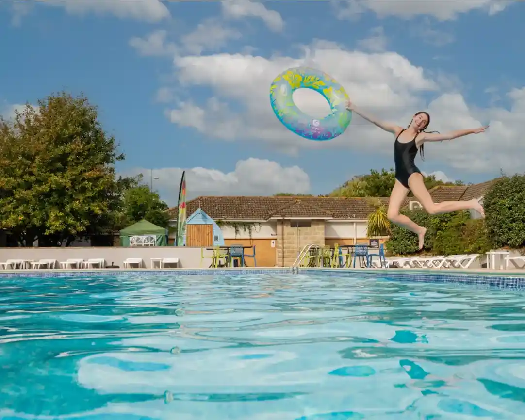 A person in a black swimsuit jumps into a clear swimming pool while holding a colorful inflatable ring. Lush greenery and a building are visible in the background under a blue sky with fluffy clouds.