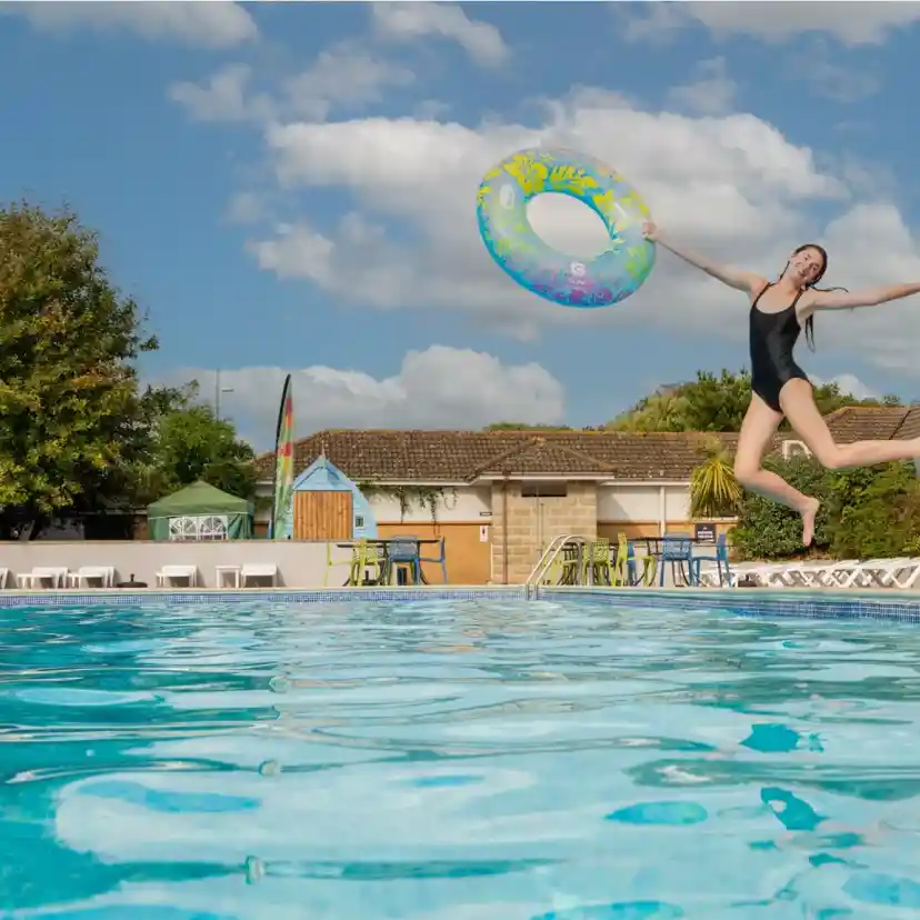 A person in a black swimsuit jumps into a clear swimming pool while holding a colorful inflatable ring. Lush greenery and a building are visible in the background under a blue sky with fluffy clouds.