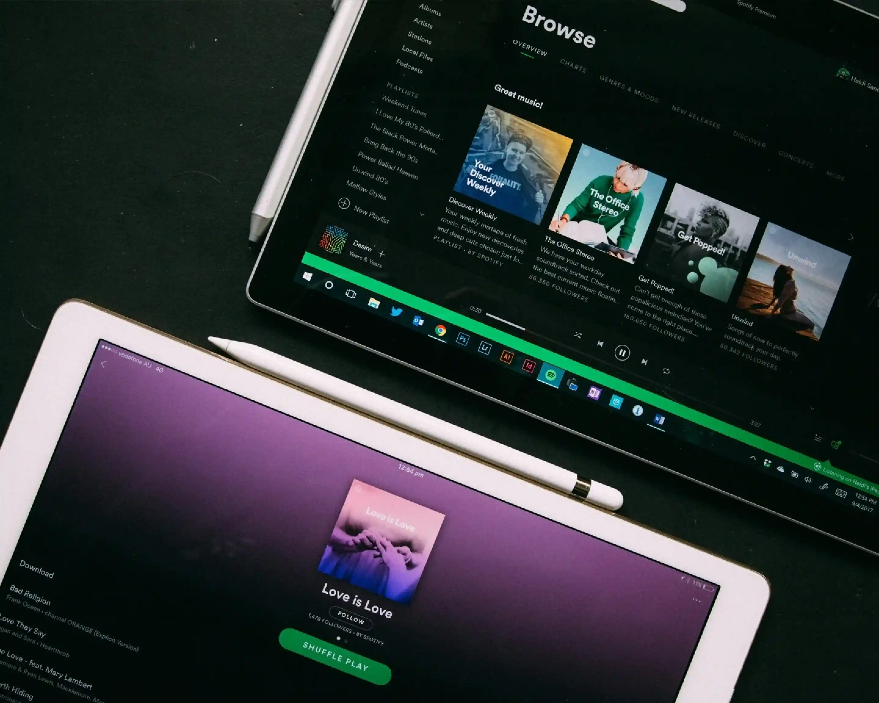 Two tablets on a dark surface, both displaying the Spotify app. The top device shows the “Browse” section with music playlists, while the bottom device shows a playlist titled “Love is Love.” Each tablet has a stylus placed nearby.