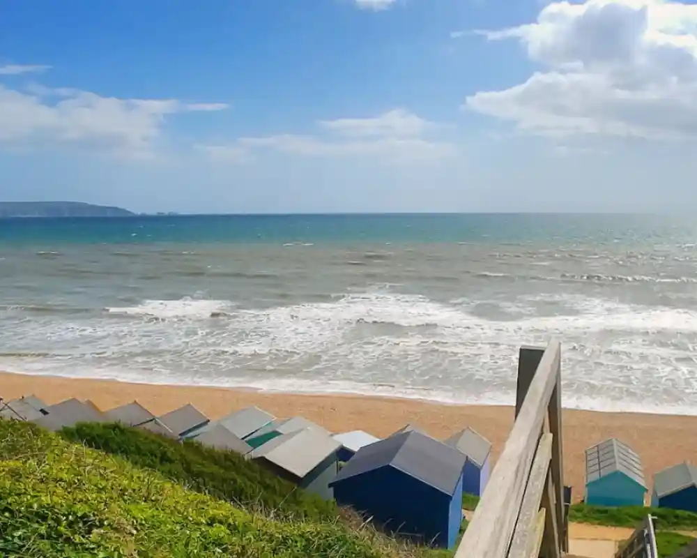 A view of a beach with rolling waves and a sandy shore, lined with colorful beach huts. Overcast skies create a serene atmosphere.
