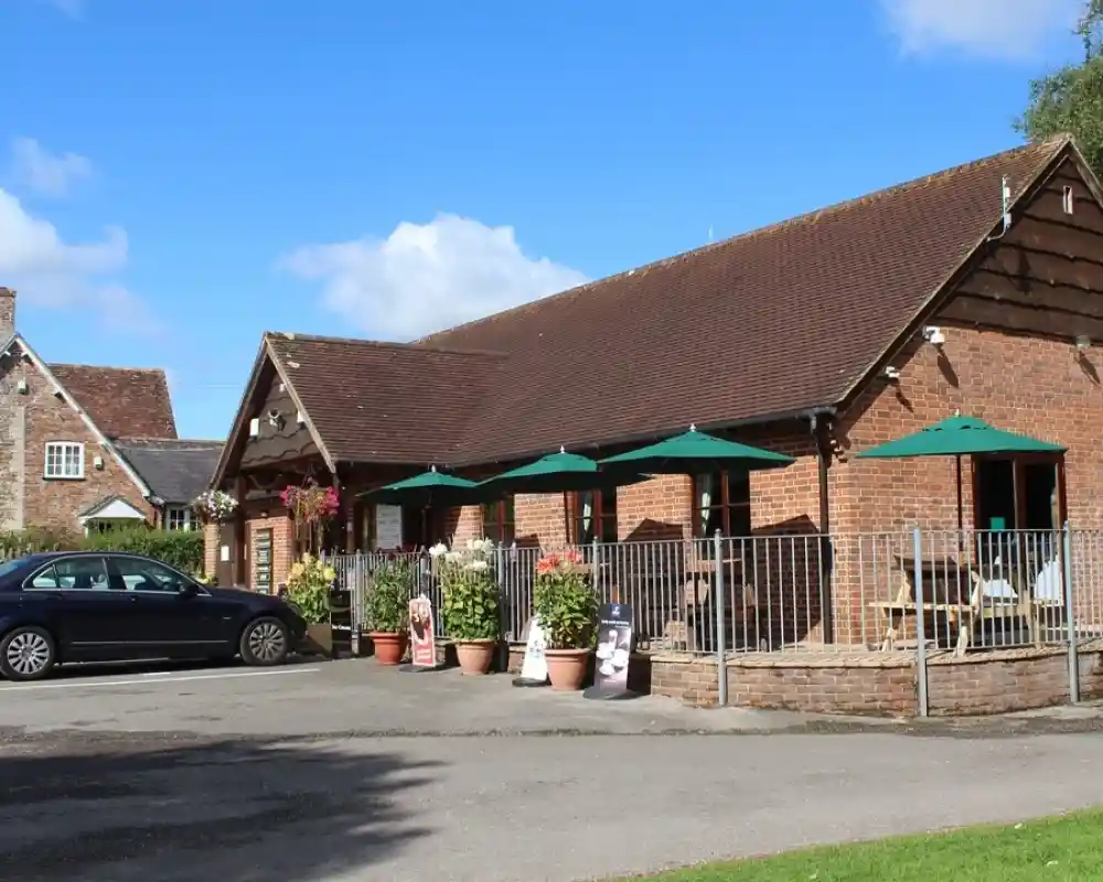 A charming brick building with a sloped roof and outdoor seating under green umbrellas. Several potted plants adorn the area, and a black car is parked nearby. Blue skies and a few clouds create a pleasant backdrop.