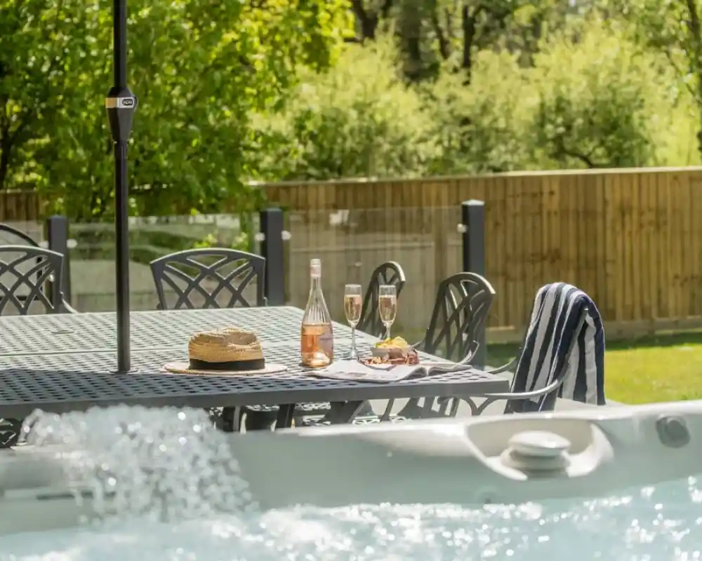 A luxurious outdoor setting features a bubbling hot tub in the foreground, with a dining table and chairs nearby. On the table, there is a bottle of rosé wine, two flutes filled with sparkling wine, a straw hat, and a small plate of snacks. Lush greenery and a wooden fence serve as the backdrop, creating a serene atmosphere.