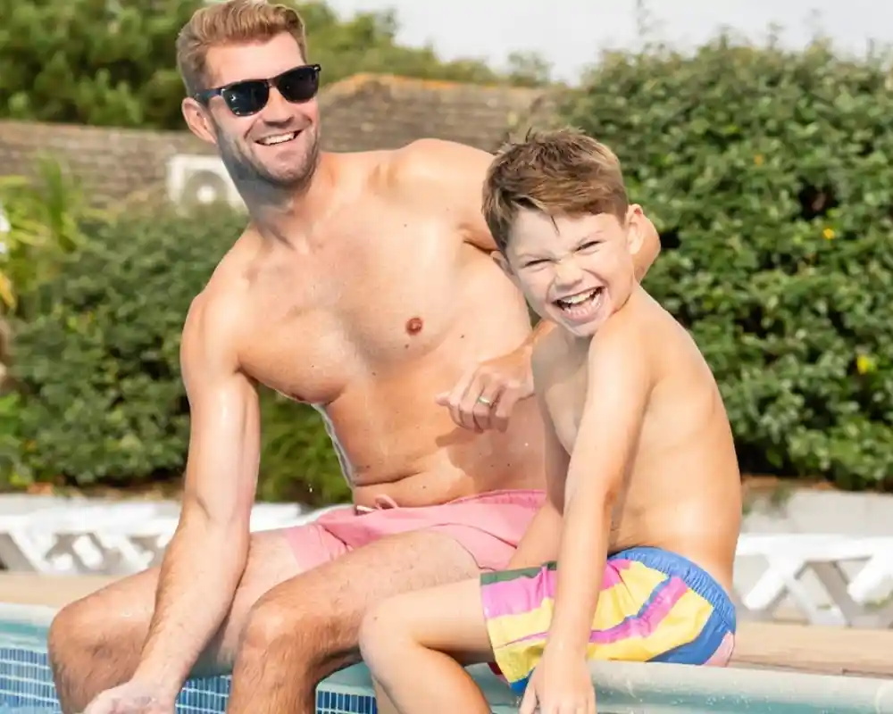 A shirtless man wearing sunglasses sits next to a smiling young boy, both enjoying a sunny day by a pool. The boy is playfully splashing water while the man laughs, surrounded by greenery.