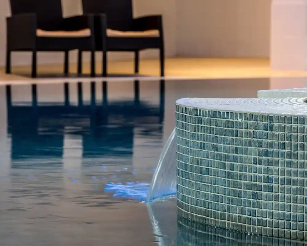 A modern, tiled poolside feature with a gentle water flow, reflecting the blue water of the pool. In the background, two black chairs are visible, adding to the tranquil atmosphere.