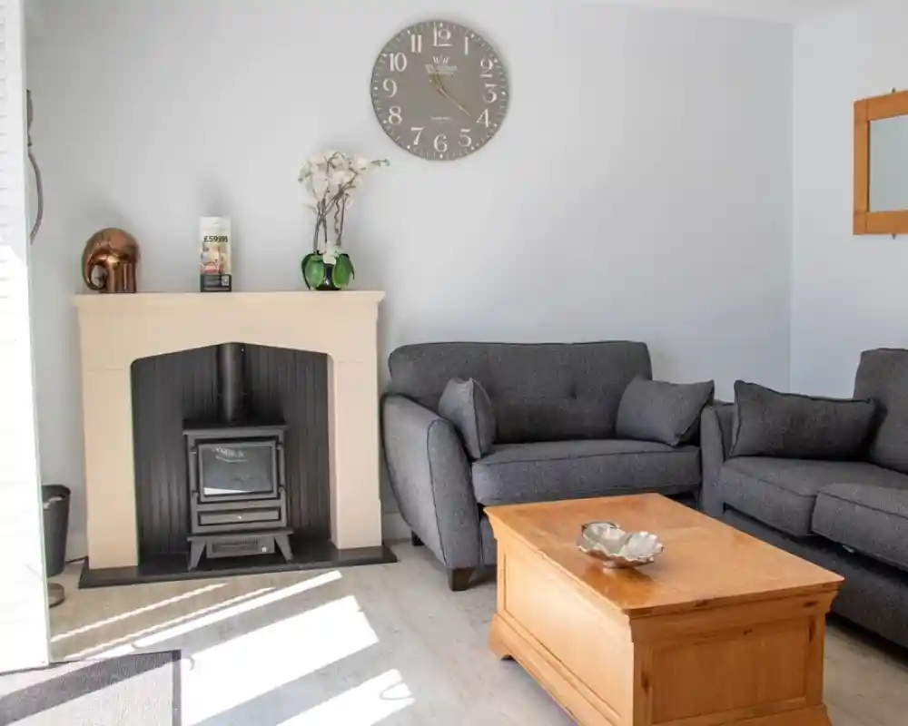 A cozy living room featuring two dark gray sofas, a wooden coffee table, and a fireplace with a modern design. A large wall clock hangs above the fireplace, and a mirror is mounted on the wall. Natural light streams in, creating a welcoming atmosphere.