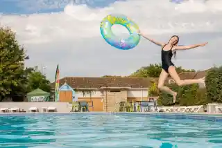 A young girl wearing a black swimsuit joyfully leaps into a swimming pool, holding a colorful inflatable ring. Lush greenery and a building can be seen in the background under a partly cloudy sky.