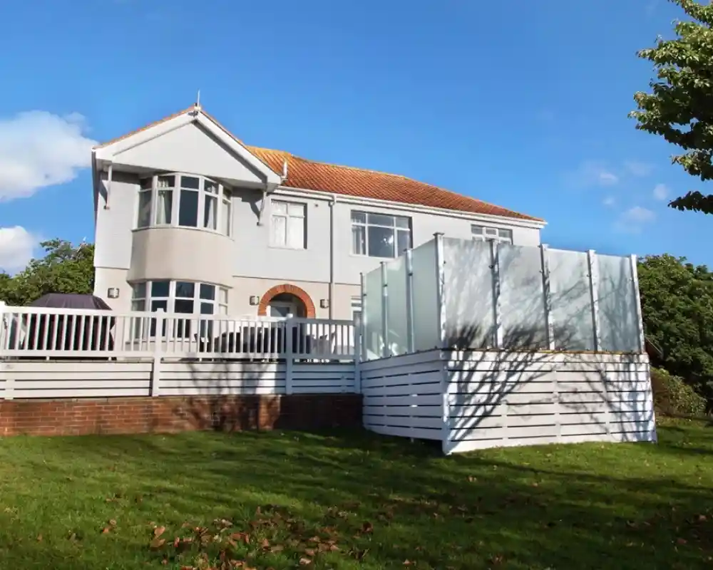 A large, modern house with a light exterior and a sloped roof. The house features a spacious deck with white railings, surrounded by green grass and trees. A glass-enclosed section is visible on the side, highlighting a contemporary design. The sky is blue with a few fluffy clouds.