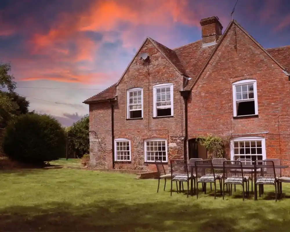 A charming brick house with multiple windows, surrounded by a lush green lawn. There are several outdoor chairs arranged in front of the house. The sky is filled with vibrant orange and purple hues during sunset.