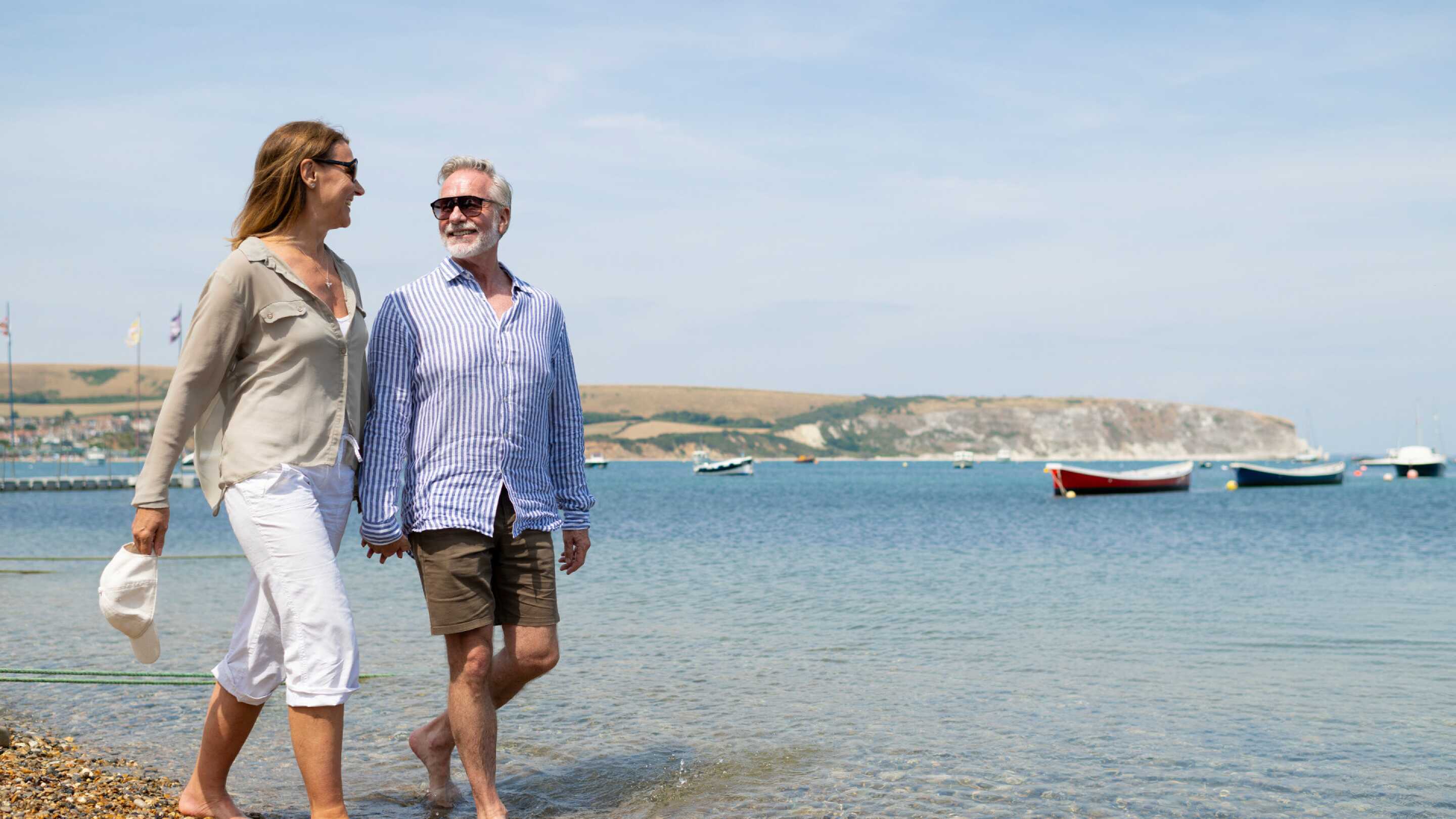 A couple walks along the shoreline, smiling at each other. The woman wears a light blouse and white pants, while the man is in a striped shirt and shorts. They stroll near gentle waves with boats anchored in the distance and a scenic backdrop of hills.