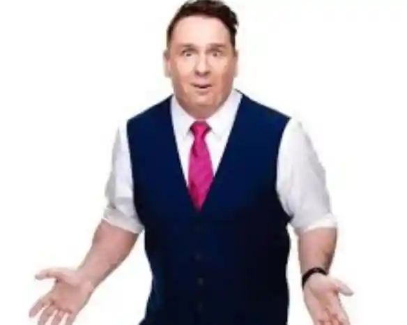 A man in a blue vest and white shirt with a pink tie stands with an expressive face, hands raised and open, conveying surprise or confusion.