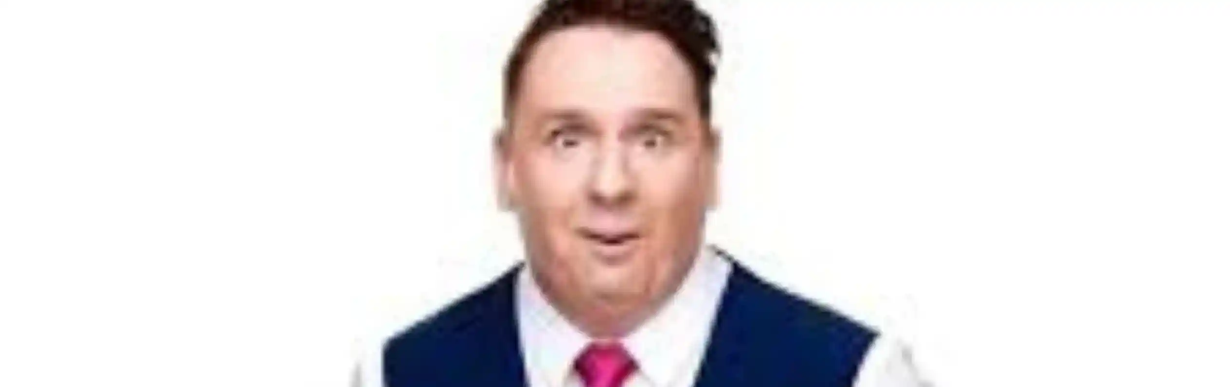 A man in a blue vest and white shirt with a pink tie stands with an expressive face, hands raised and open, conveying surprise or confusion.