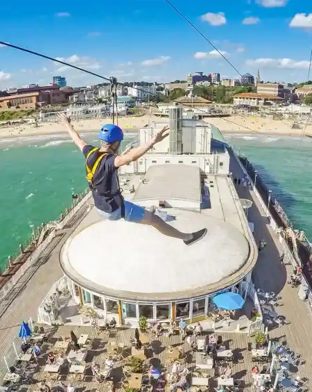 A person enjoys a zip line ride over a beachside pier, arms outstretched, with a vibrant blue sky and green ocean visible below. The pier features outdoor seating and people enjoying the sun.