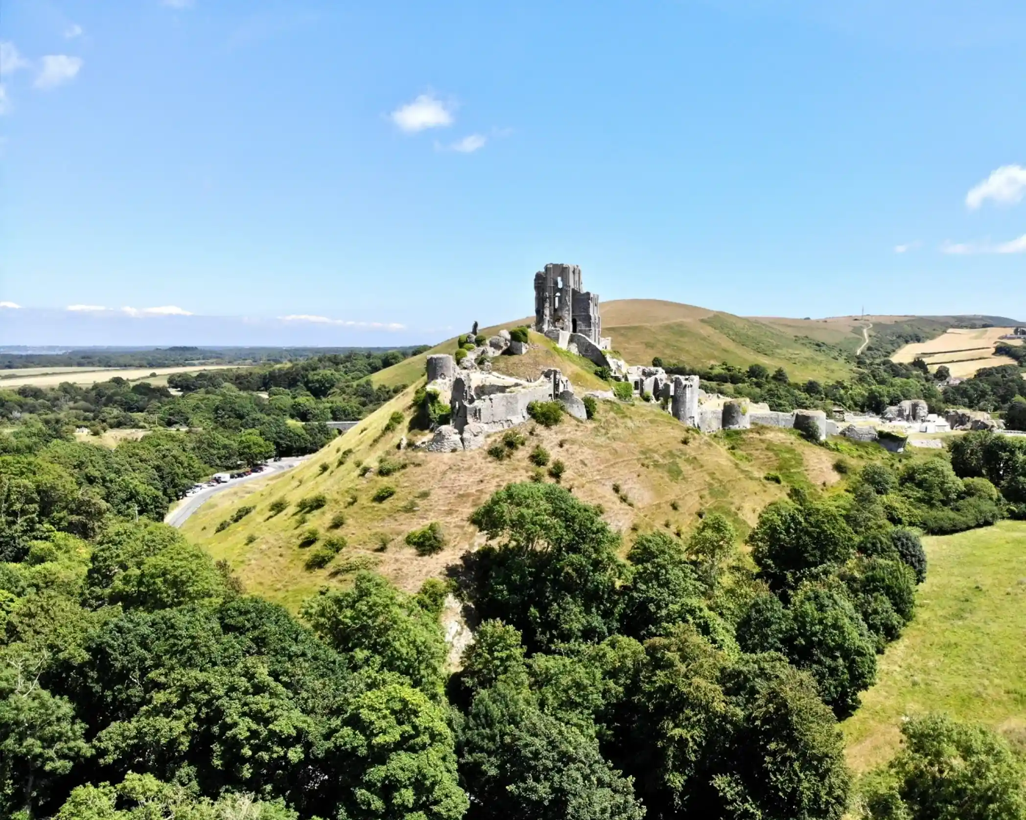 Aerial view of medieval ruins on a hillside, surrounded by lush greenery and rolling hills under a clear blue sky.