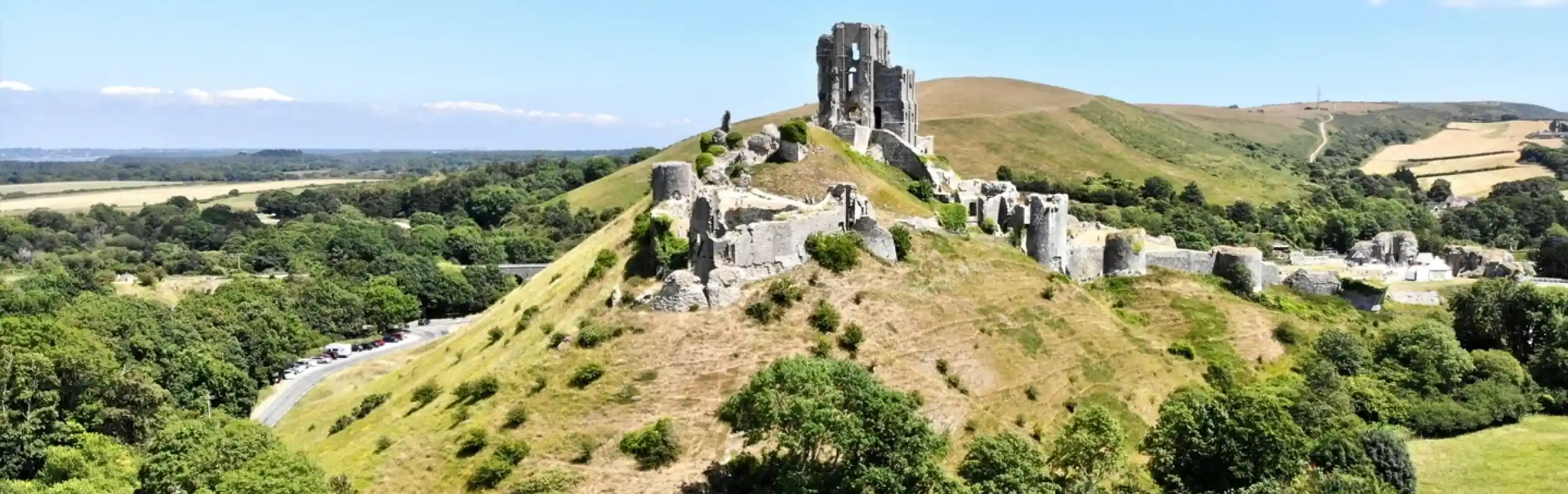 Aerial view of medieval ruins on a hillside, surrounded by lush greenery and rolling hills under a clear blue sky.