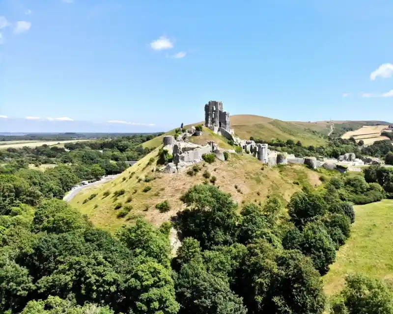 Aerial view of medieval ruins on a hillside, surrounded by lush greenery and rolling hills under a clear blue sky.