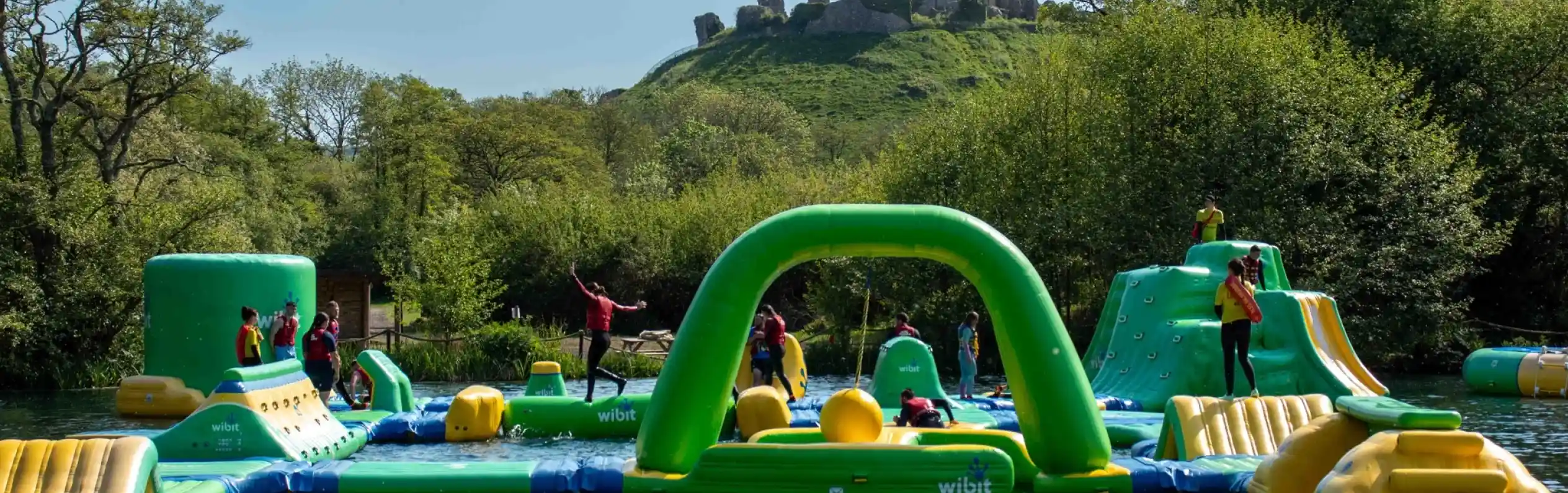 A vibrant inflatable water park is set up on a lake, with people actively engaging in various activities on the structures. In the background, a rocky hill topped with ruins is visible, surrounded by lush greenery under a clear blue sky.