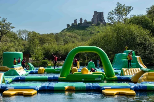 A vibrant inflatable water park is set up on a lake, with people actively engaging in various activities on the structures. In the background, a rocky hill topped with ruins is visible, surrounded by lush greenery under a clear blue sky.