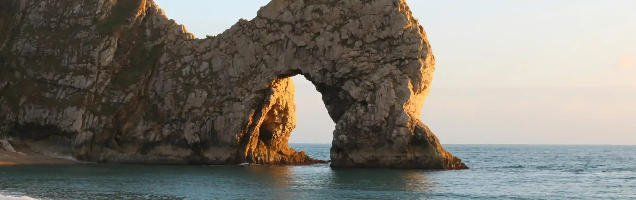 A sunlit rock formation resembling an arch stands in the ocean, with waves gently lapping at the sandy beach in the foreground. The sky is clear, casting warm colors on the cliffs and rock.