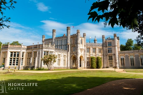 Highcliffe Castle with intricate architecture and large windows, set against a bright blue sky and green lawns. The castle features towers and is surrounded by trees.