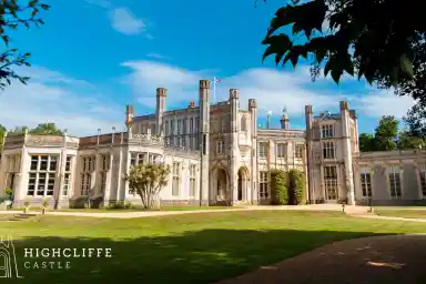 Highcliffe Castle with intricate architecture and large windows, set against a bright blue sky and green lawns. The castle features towers and is surrounded by trees.