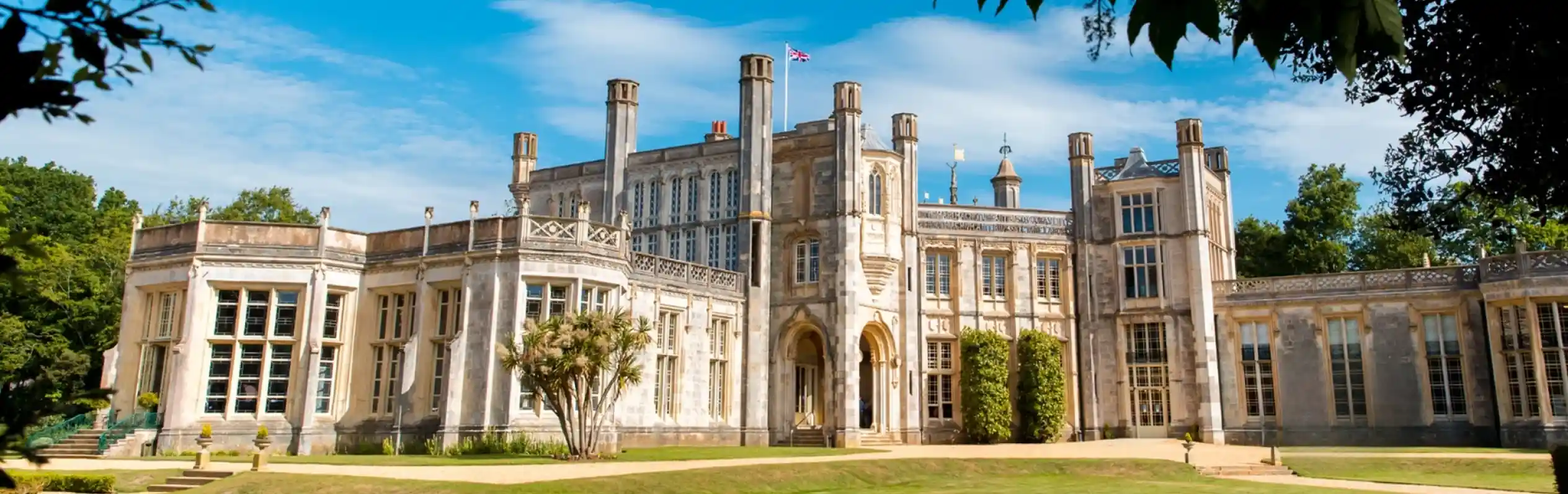 Highcliffe Castle with intricate architecture and large windows, set against a bright blue sky and green lawns. The castle features towers and is surrounded by trees.