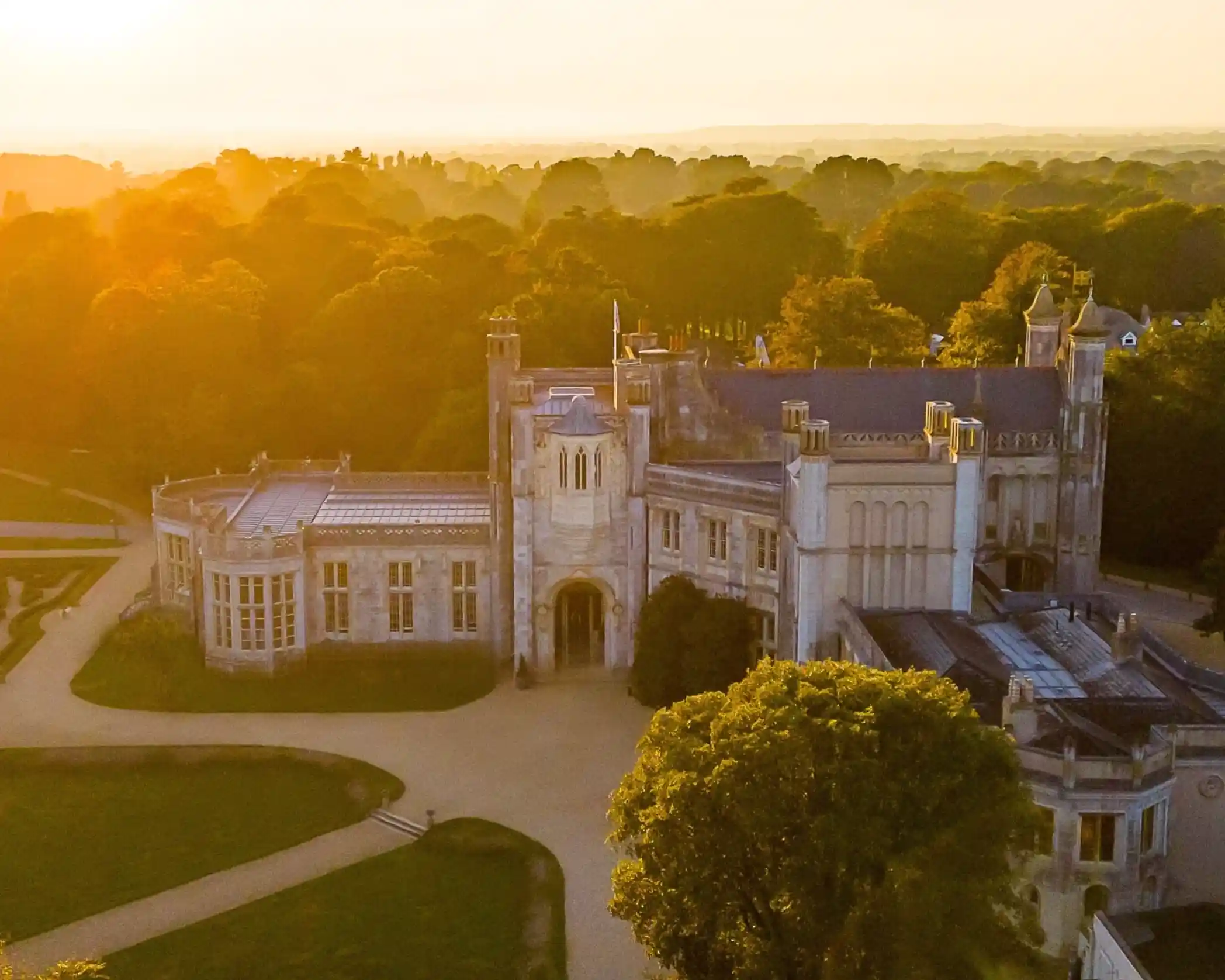 A grand historic mansion surrounded by lush trees, illuminated by a warm golden sunset. The architecture features turrets and detailed stonework, with pathways winding around the property. Photo credit -Andrew Millard