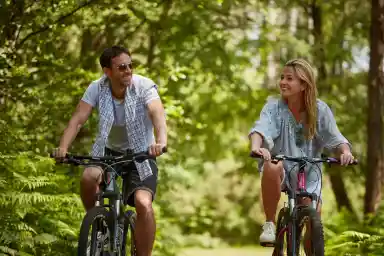 A man and woman ride bicycles along a wooded path, smiling at each other. Lush green foliage surrounds them, creating a serene outdoor atmosphere.