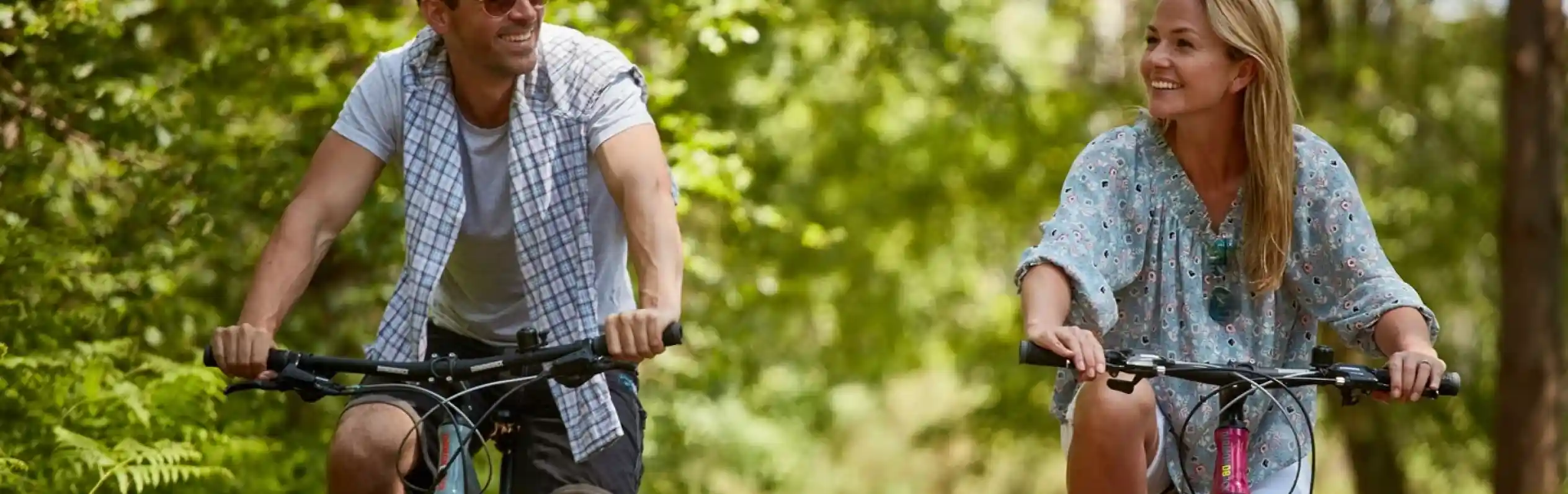 A man and woman ride bicycles along a wooded path, smiling at each other. Lush green foliage surrounds them, creating a serene outdoor atmosphere.
