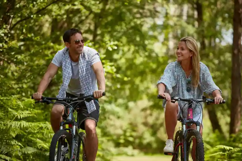 A man and woman ride bicycles along a wooded path, smiling at each other. Lush green foliage surrounds them, creating a serene outdoor atmosphere.