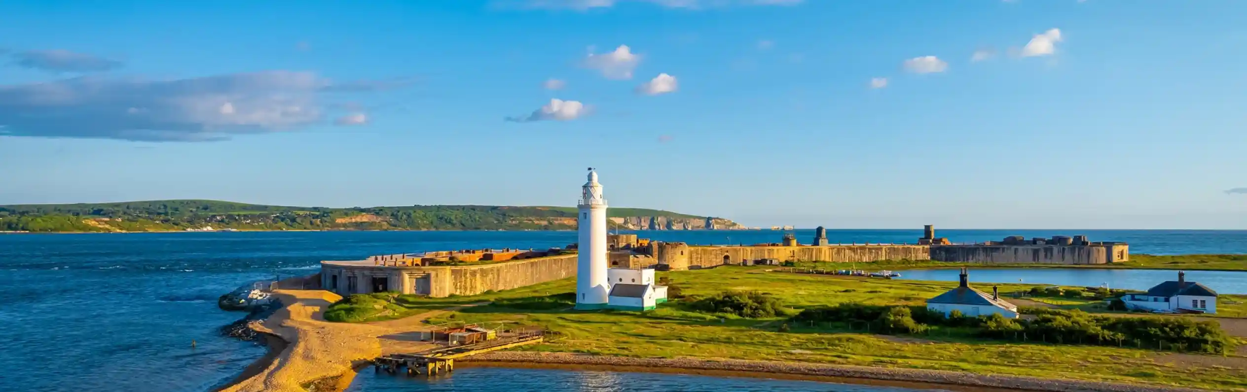 A coastal scene featuring a white lighthouse on a grassy shore, surrounded by a stone fortification and calm blue waters. Clear skies with scattered clouds and a distant green hillside add to the picturesque landscape.