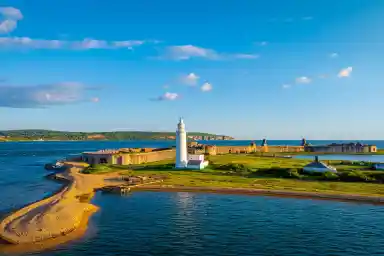 A coastal scene featuring a white lighthouse on a grassy shore, surrounded by a stone fortification and calm blue waters. Clear skies with scattered clouds and a distant green hillside add to the picturesque landscape.