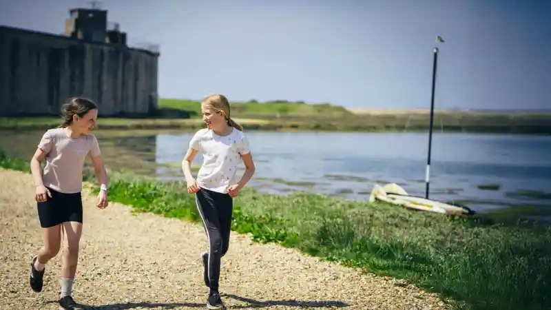 Two girls run along a gravel path near a calm shoreline. In the background, a sailboat is anchored, and a large stone structure stands beside them. It's a bright and sunny day.