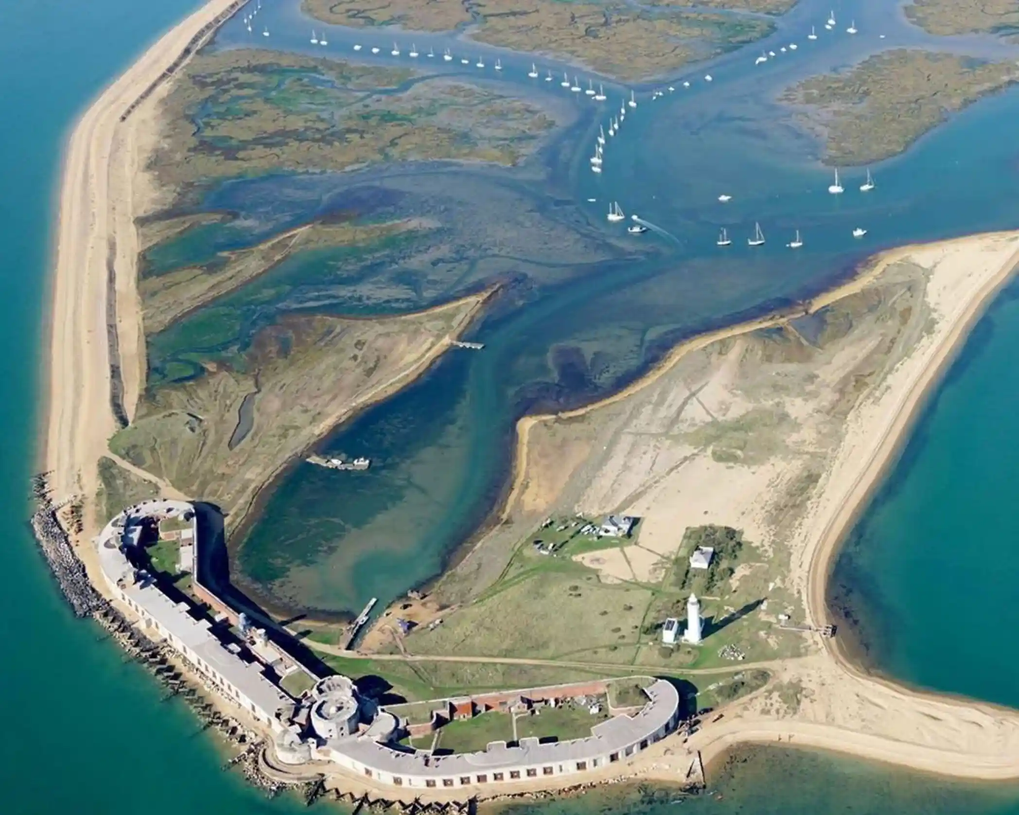 Aerial view of a coastal island featuring a historic fortification, sandy beaches, and patches of greenery. Sailboats dot the calm waters surrounding the island.