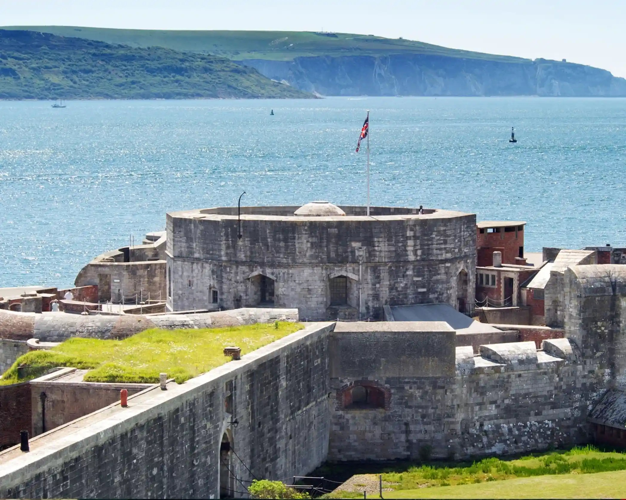 A historic stone fort with a circular tower and a flag flying at the top overlooks a calm blue sea. In the background, green hills rise against a clear sky, dotted with small boats.