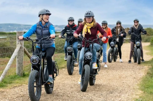 A group of seven people riding electric bikes on a gravel path, smiling and chatting. They are wearing helmets and casual clothing, with scenic hills in the background.