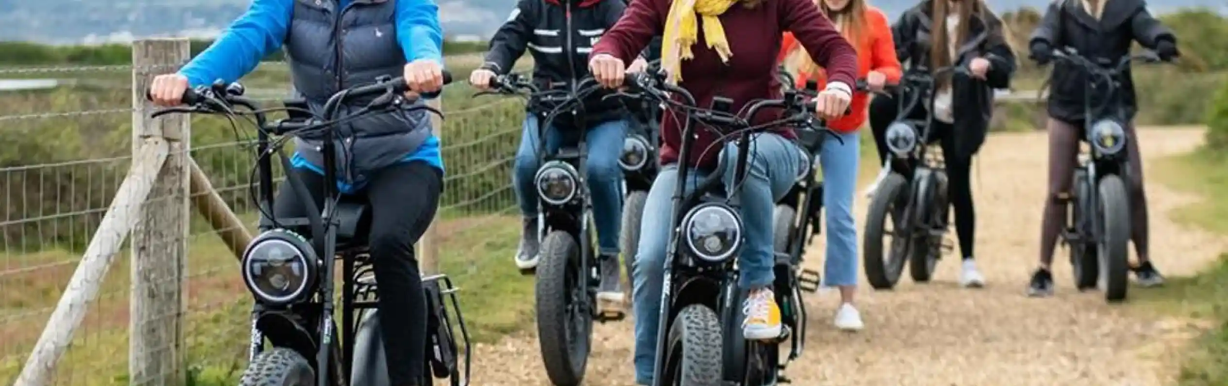 A group of seven people riding electric bikes on a gravel path, smiling and chatting. They are wearing helmets and casual clothing, with scenic hills in the background.