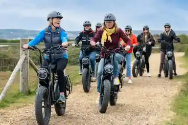 A group of seven people riding electric bikes on a gravel path, smiling and chatting. They are wearing helmets and casual clothing, with scenic hills in the background.